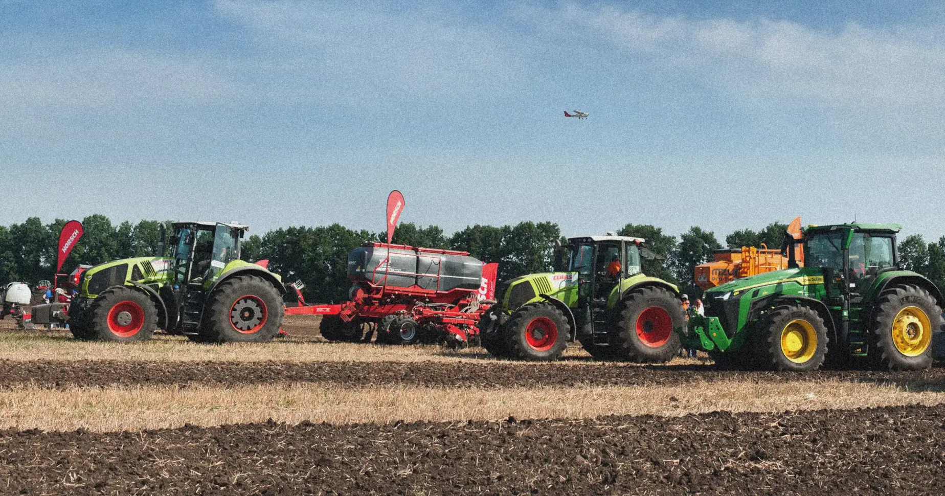 FL-How-Hidden-Fuel-Costs-Are-Slowing-Landscaping-Fleets - Fuel Logic Line of green and yellow tractors with red wheels and red attached equipment in a plowed field at an agricultural show under a blue sky.